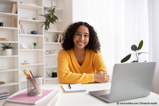 Portrait of happy young black woman sitting at desk with study materials and laptop computer, smiling at camera indoors Frau am Schreibtisch