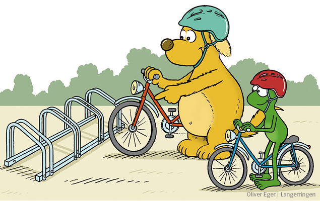 Mini und Max stellen ihre Fahrräder in den Fahrradständer. Max trägt einen grünen Helm, Minis Helm ist rot.